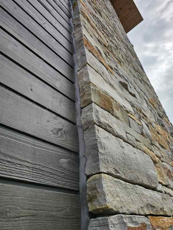 Looking closely at a completed siding expansion joint showing a clean grey commercial sealant bead bonding dark grey horizontal cladding panels to a textured stone masonry wall.