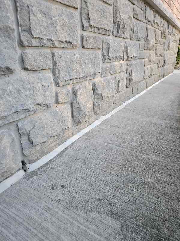 Completed exterior caulking showing a crisp grey commercial sealant bead bonding a concrete porch pad to a long, rugged split-face stone wall.