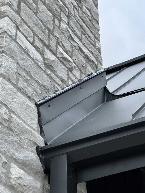 Looking up a roofline at a completed exterior transition caulking job showing a dark grey hybrid sealant bead bonding a black standing seam metal flashing track to a rugged stone masonry wall.