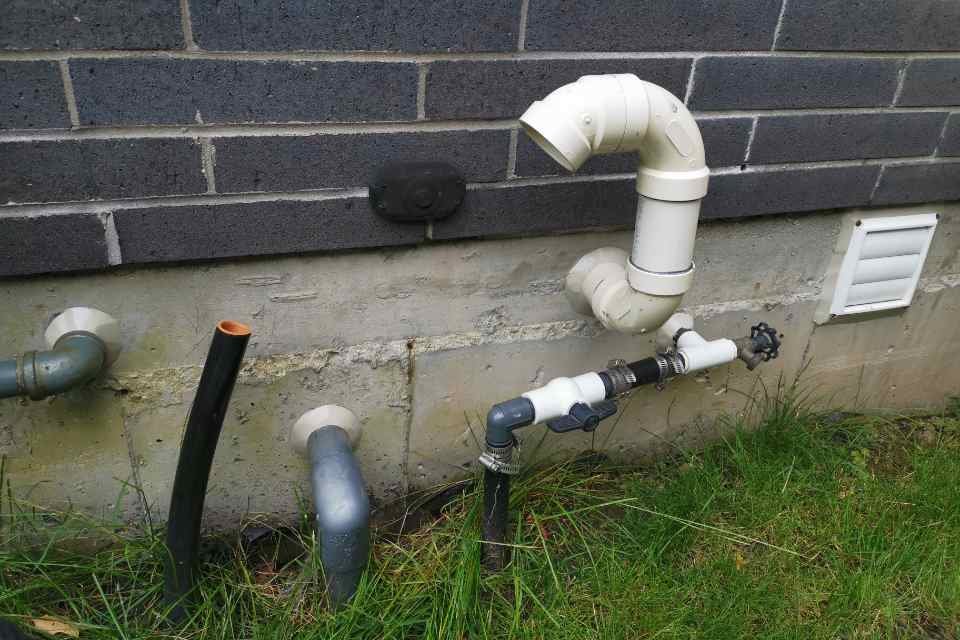A close-up view of a concrete foundation wall showing complex utility penetrations including an outdoor spigot, PVC lines, and electrical conduits requiring exterior utility caulking.
