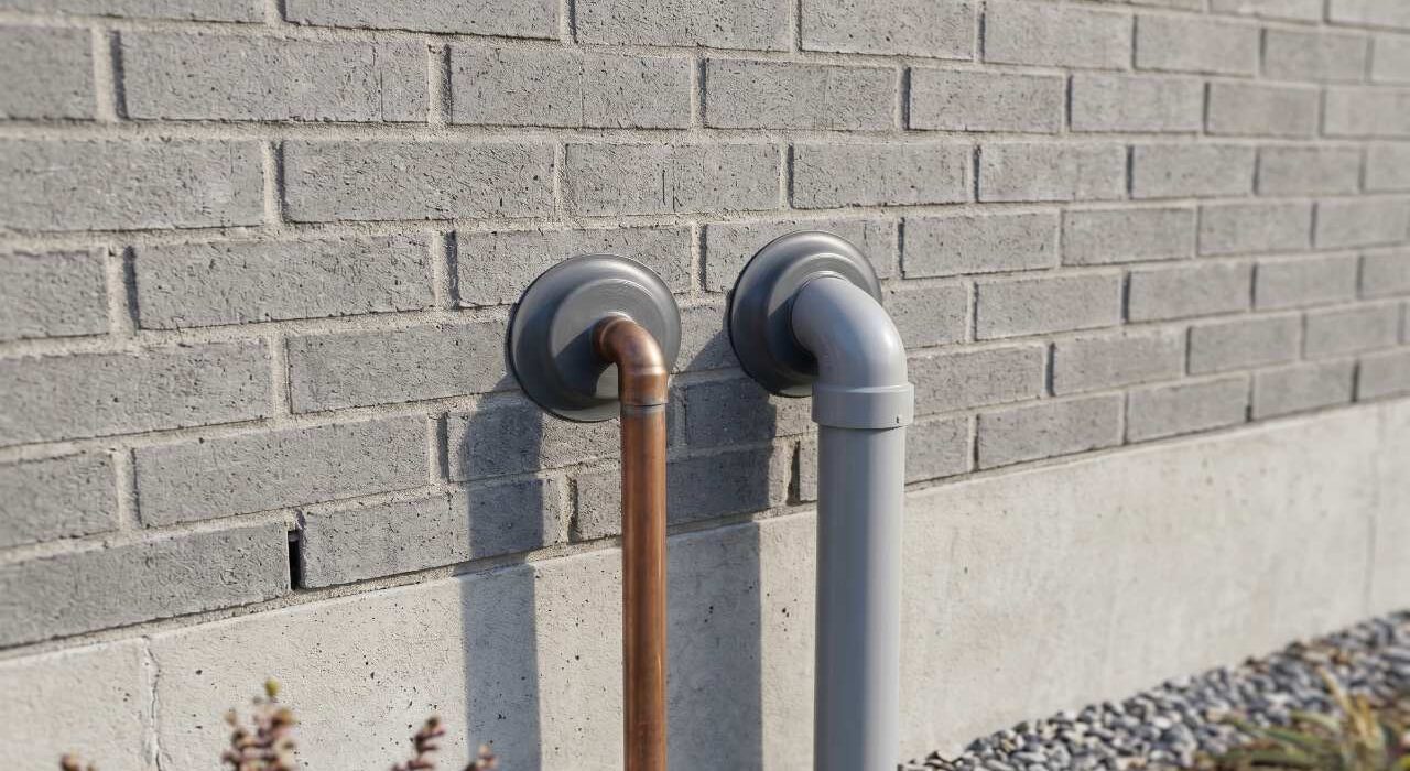 A close-up view of an exterior grey brick wall showing a copper line and grey PVC pipe with plastic escutcheon plates hiding the underlying utility caulking.