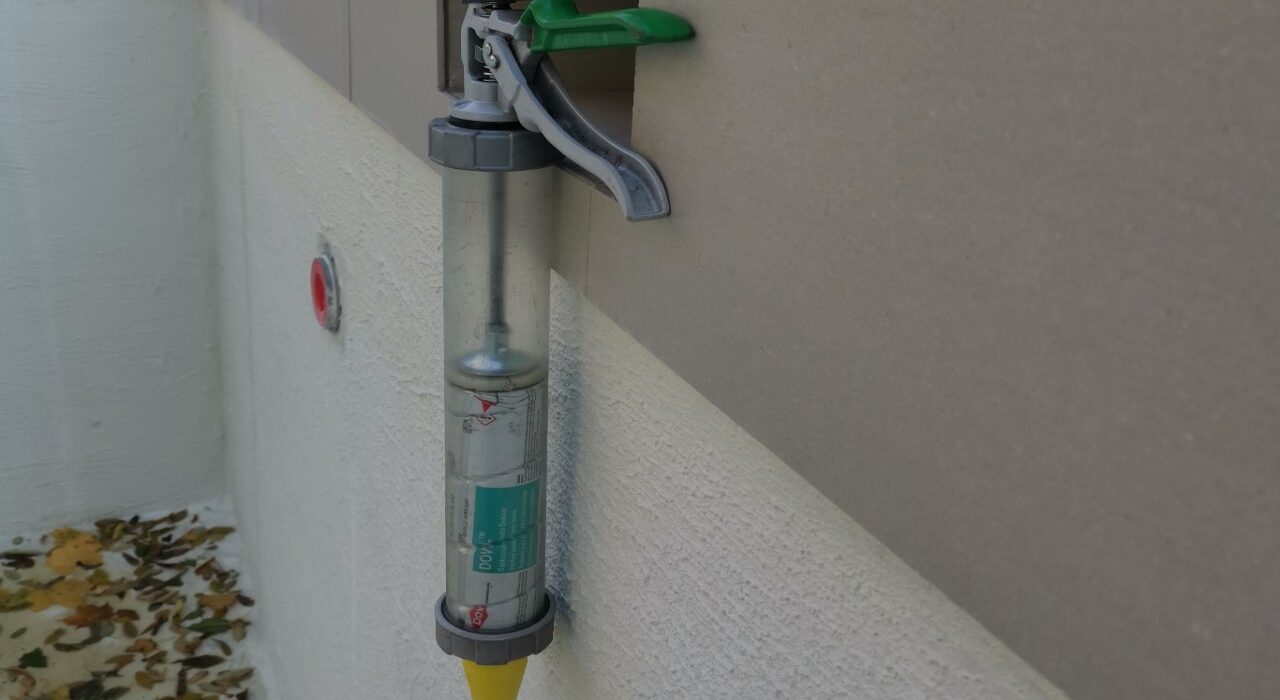 A professional clear-barrel Albion sausage caulking gun with a high-thrust green trigger frame hanging on a pool deck coping stone joint.
