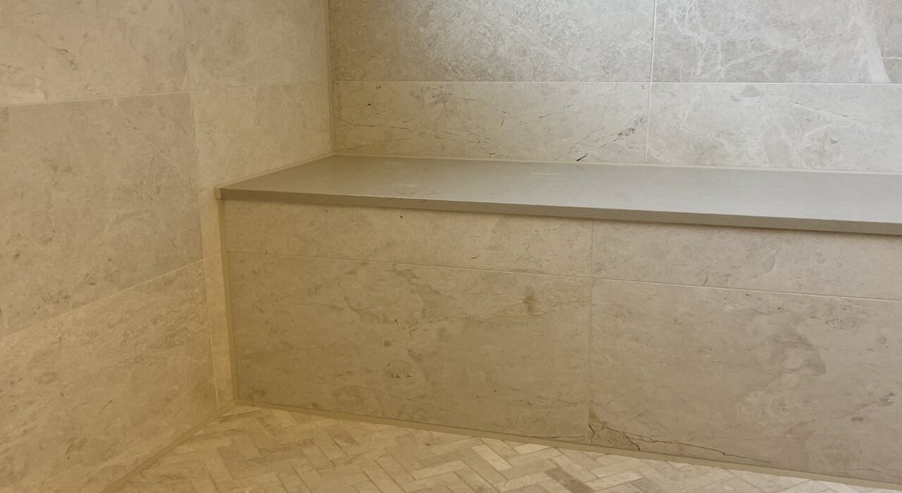 A thin, perfectly tooled neutral-cure silicone joint applied cleanly along the marble tile walls, herringbone floor, and stone bench of a premium residential shower.
