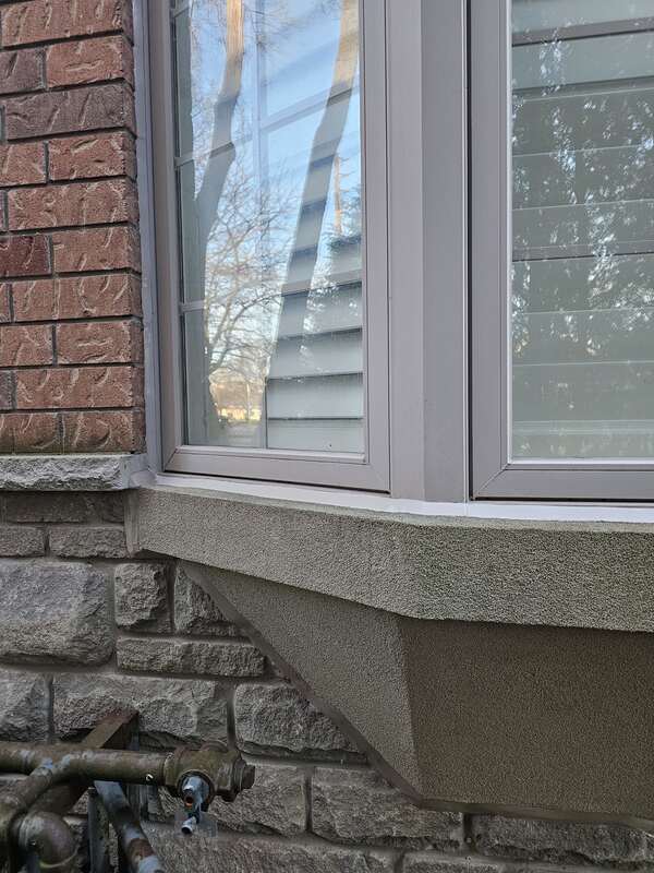 A close-up side view of a brick house showing fresh white caulking sealing a bay window frame to a concrete sill, brick wall, and natural stone veneer.