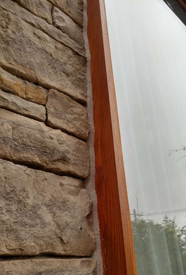 A textured, sandy-colored masonry hybrid sealant applied flawlessly along a rugged natural ledger stone wall and stained cedar wood window frame casing.