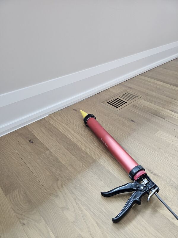 A close-up view of an interior room showing a professional red caulking gun resting on oak hardwood floorboards next to a white baseboard.