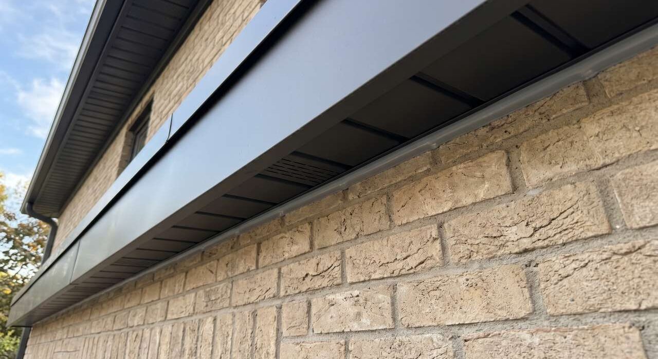 Looking up at a long horizontal black aluminum eave soffit panel flawlessly caulked against a modern residential brick wall facade.