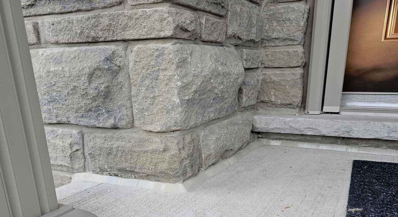 A close-up view of a completed concrete porch caulking job showing a flawless beige polymer bead sealing a poured concrete landing pad to a rugged stone masonry pillar wall.