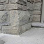A close-up view of a completed concrete porch caulking job showing a flawless beige polymer bead sealing a poured concrete landing pad to a rugged stone masonry pillar wall.