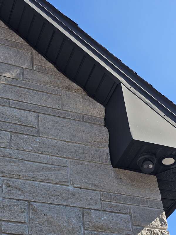 Macro view of a completed exterior soffit caulking joint where black aluminum roof trim meets a split-face stone wall with a tooled weatherseal.