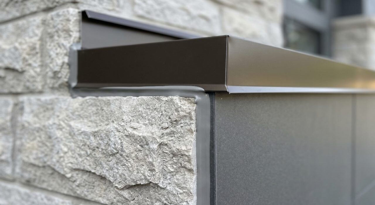 A close-up view of a completed exterior transition caulking job showing a flawless grey hybrid sealant bead sealing a dark bronze aluminum flashing track to a rugged stone masonry wall.