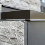 A close-up view of a completed exterior transition caulking job showing a flawless grey hybrid sealant bead sealing a dark bronze aluminum flashing track to a rugged stone masonry wall.