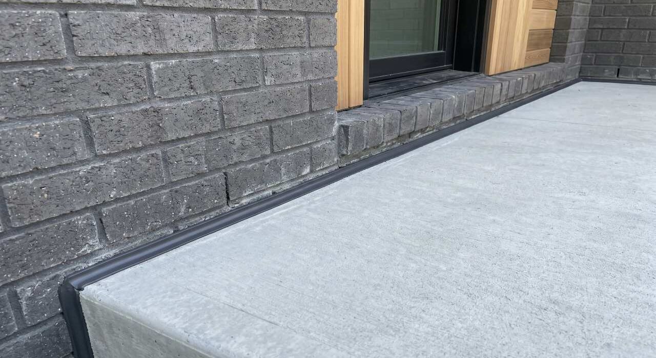 A close-up view of a completed porch to house expansion joint caulking job showing a flawless charcoal polyurethane bead sealing a concrete porch step to a grey brick wall.