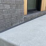 A close-up view of a completed porch to house expansion joint caulking job showing a flawless charcoal polyurethane bead sealing a concrete porch step to a grey brick wall.