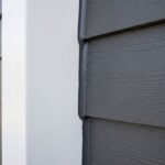 A close-up view of a completed siding and trim caulking job showing a flawless charcoal polymer bead sealing dark grey fibre cement siding planks to a white vertical corner trim board.