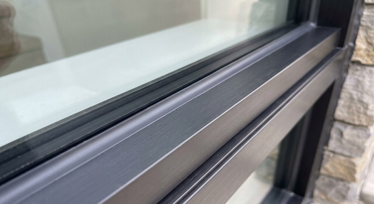 A close-up 4:3 view of a completed window glazing caulking job showing a flawless grey silicone bead sealing an insulated glass pane to a dark aluminum frame sash.