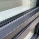 A close-up 4:3 view of a completed window glazing caulking job showing a flawless grey silicone bead sealing an insulated glass pane to a dark aluminum frame sash.
