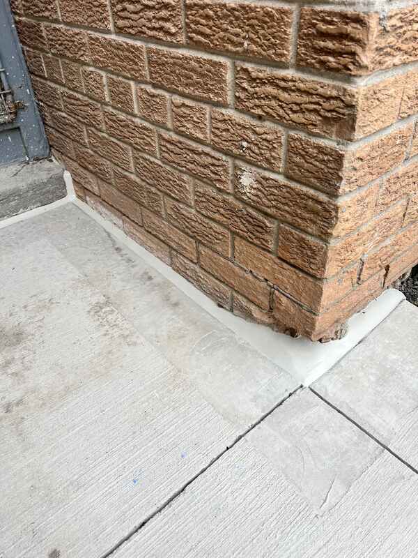 A heavy-duty, perfectly tooled grey polyurethane sealant bead applied flawlessly along a horizontal concrete sidewalk and vertical red brick foundation wall corner.