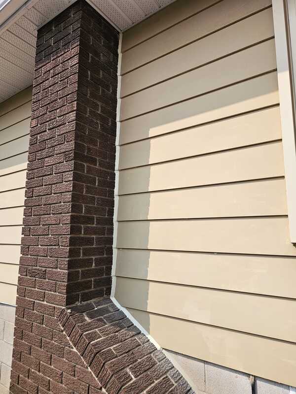 Completed exterior caulking showing a crisp white commercial sealant bead bonding tan horizontal lap siding boards to a dark brick chimney wall.