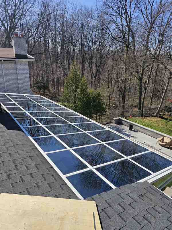 Looking down at a massive completed multi-panel glass sunroom roof integration on an asphalt shingle roofline showing white structural frame weatherseals.