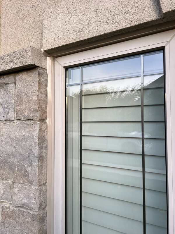 A close-up view of a completed exterior window caulking job on a house showing clean sealant lines transitioning between stucco, stone veneer, and vinyl frames.