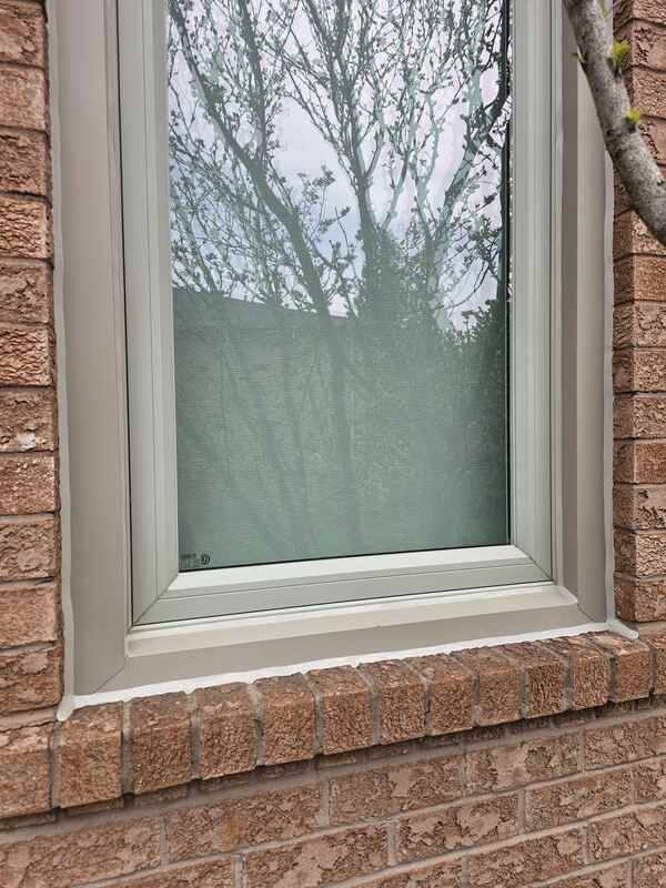 A tight vertical close-up view of an exterior brick wall showing fresh white caulking sealing a light-grey window frame to a sloped rowlock brick sill.