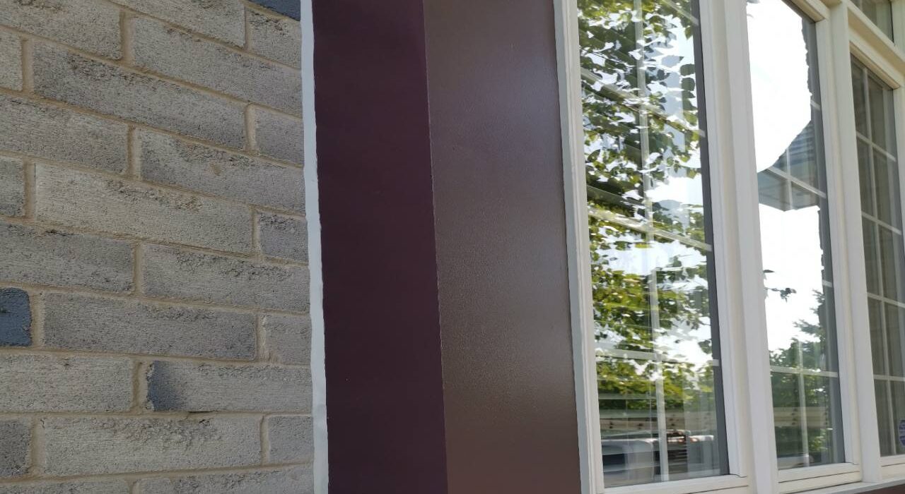 A crisp vertical white elastomeric sealant joint applied flawlessly into the mortar gaps of a grey brick wall and a smooth brown wood window trim casing.