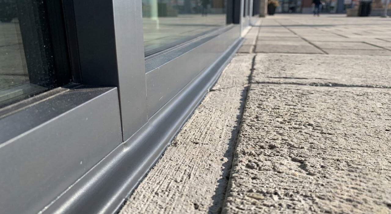 A close-up photograph of a professional commercial storefront caulking job sealing a horizontal apron expansion joint next to a retail concrete sidewalk.